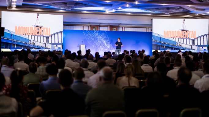 Conference coverage Wide view of a conference audience watching a speaker on a blue-lit stage